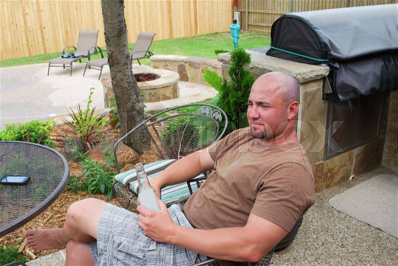 Cute bald muticultural man sitting on a patio in the back yard by bbq grill near his cell phone drinking a beer, stock photo
