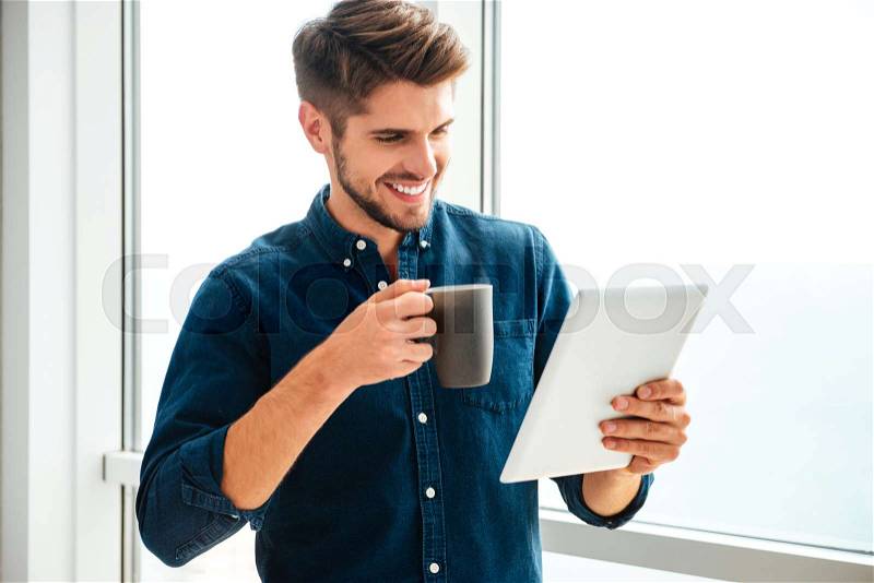 Photo of young happy man holding tablet and drinking coffee near window. Looking at tablet, stock photo