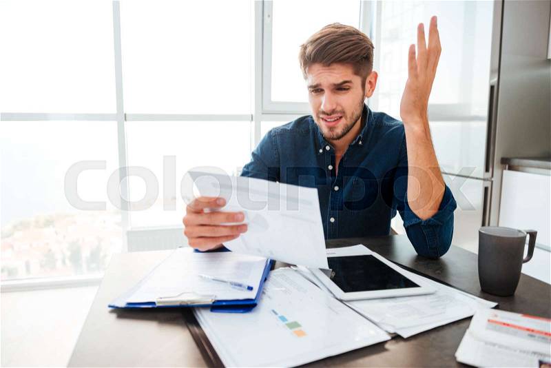 Young sad man analyzing home finances while gesturing with hand and looking at documents. Sitting near table with tablet, stock photo
