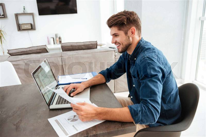 Photo of happy man using laptop and sitting at the table while looking at the papers, stock photo
