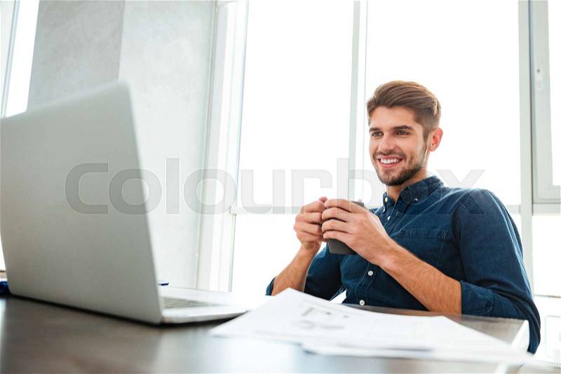 Photo of young happy man drinking tea and sitting near table with laptop and documents. Looking at laptop, stock photo