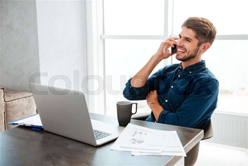 Photography of young happy man dressed in blue shirt drinking tea and sitting near table with laptop and documents while talking at his phone. Looking at laptop, stock photo