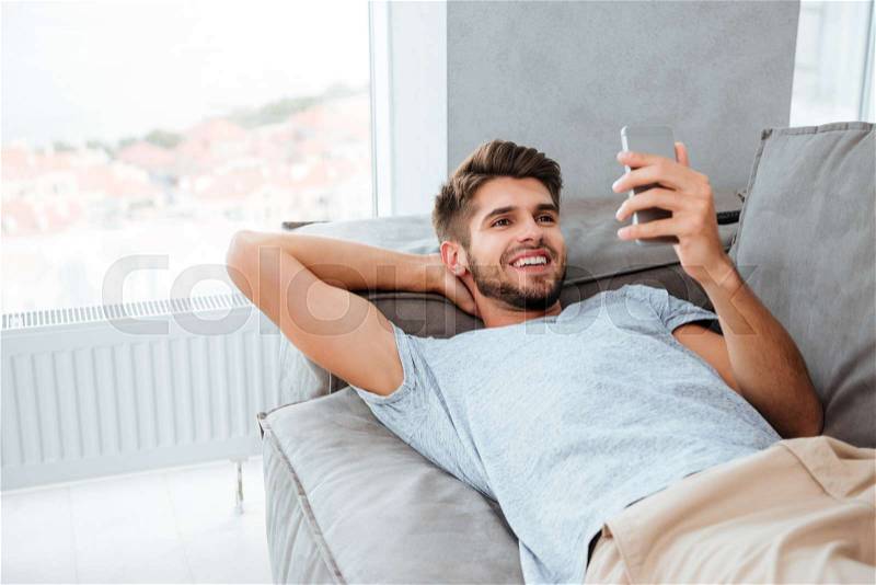 Picture of happy young man lies on bed and looking on phone, stock photo