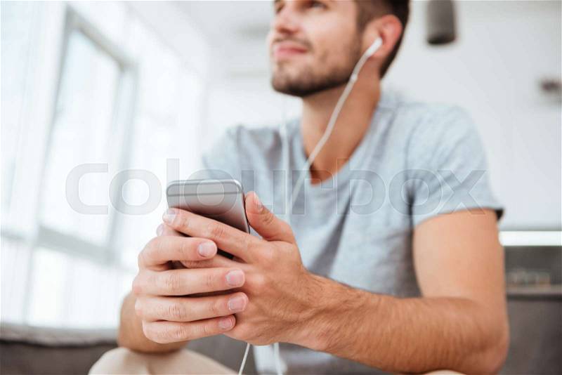 Photo of happy man dressed in t-shirt listening to music while sitting on sofa and look aside. Focus on cellphone and hands, stock photo