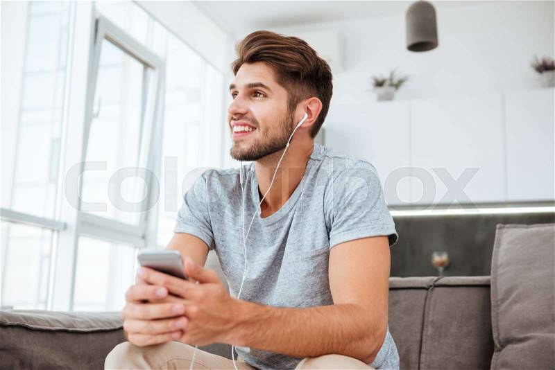 Picture of happy man dressed in t-shirt listening to music while sitting on sofa and look aside, stock photo