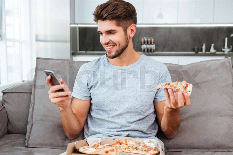 Photo of happy young man chatting and eating pizza while sitting on sofa, stock photo