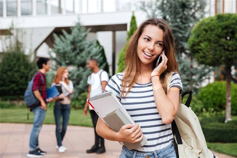 Happy lovely young woman student standing and talking on cell phone outdoors, stock photo