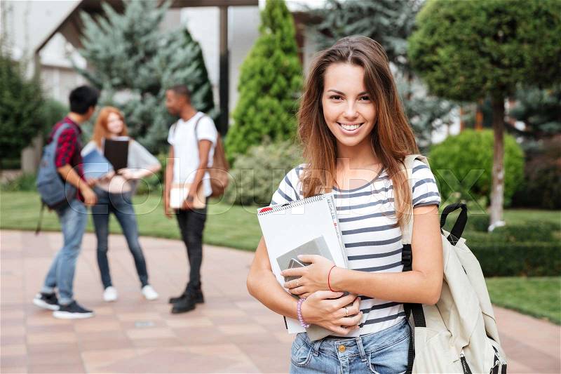 Cheerful beautiful young woman student with backpack standing in campus, stock photo