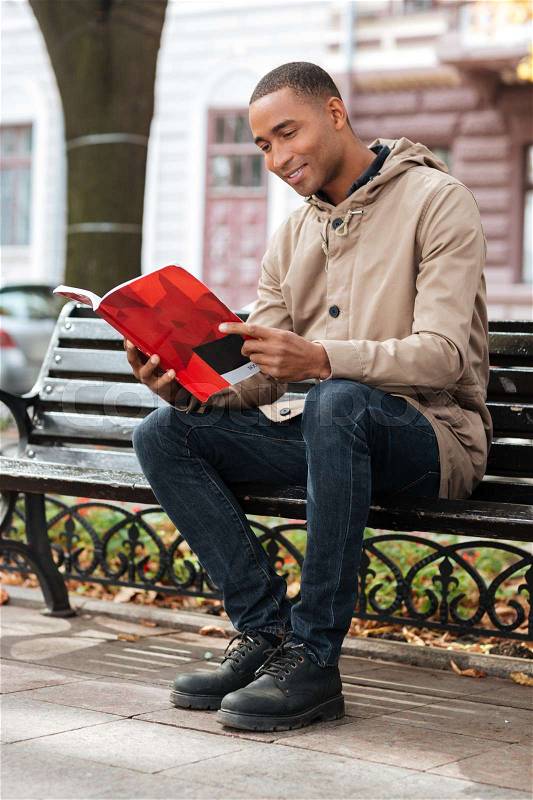 Photo of young african happy man reading a book while sitting on the bench at street, stock photo