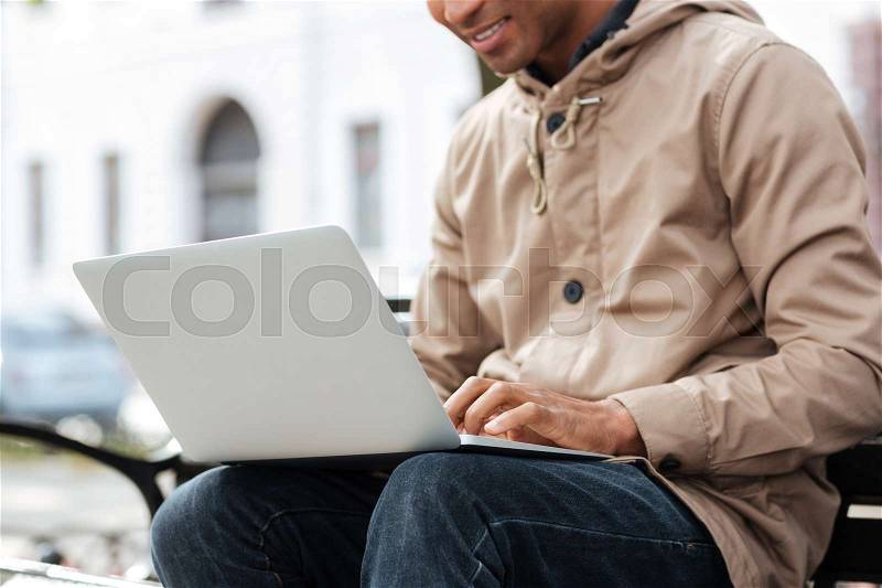 Cropped photo of young african cheerful man with laptop sitting on a wooden bench and typing, stock photo