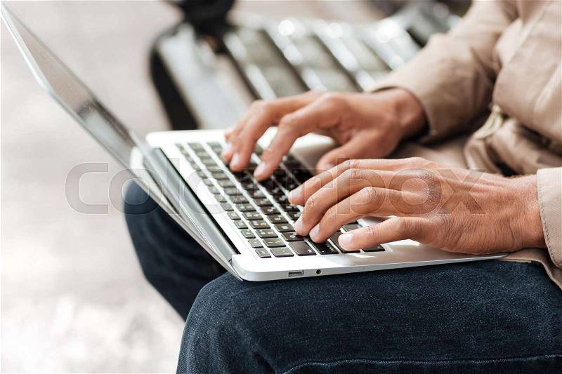 Cropped image of young african man with laptop sitting on a wooden bench and typing, stock photo