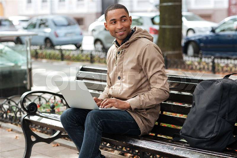 Photo of african happy man with laptop sitting on a wooden bench near backpack and typing. Look aside, stock photo