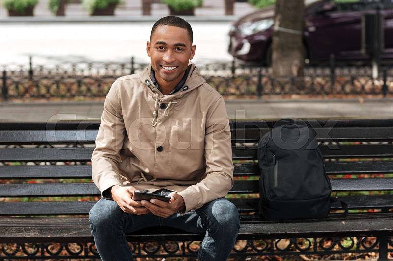 Picture of african happy man holding tablet while sitting on wooden bench near backpack and chatting. Look at camera, stock photo