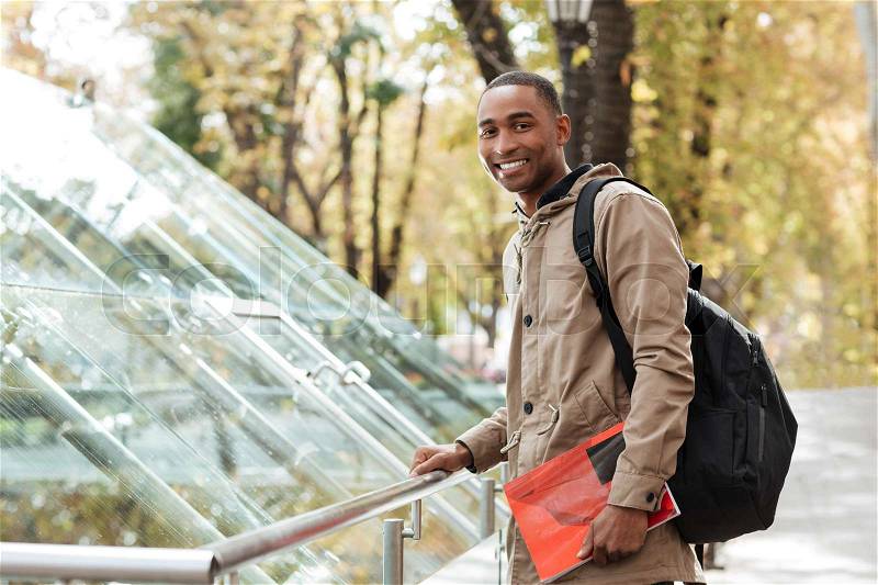 Image of young african cheerful man wearing backpack standing in the street while holding book and look at camera, stock photo
