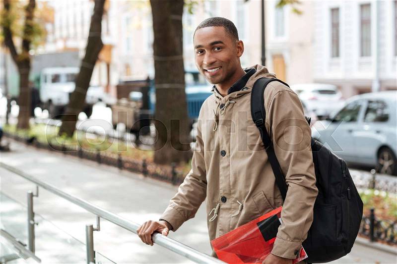 Photo of young african happy man wearing backpack standing in the street while holding book and looking at camera, stock photo