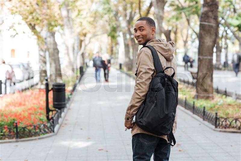 Photo of young african happy man wearing backpack walking on the street while holding book and looking at camera, stock photo