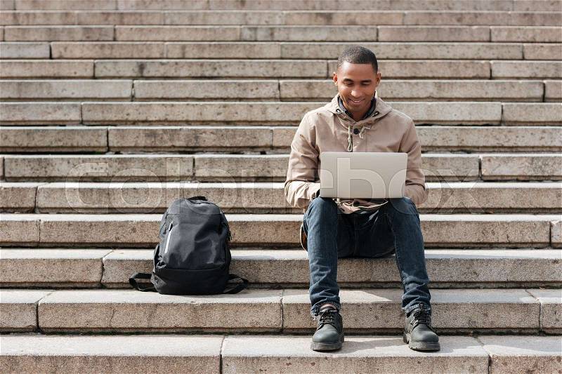 Picture of young african happy man sitting on staircase and using laptop. Looking at laptop, stock photo