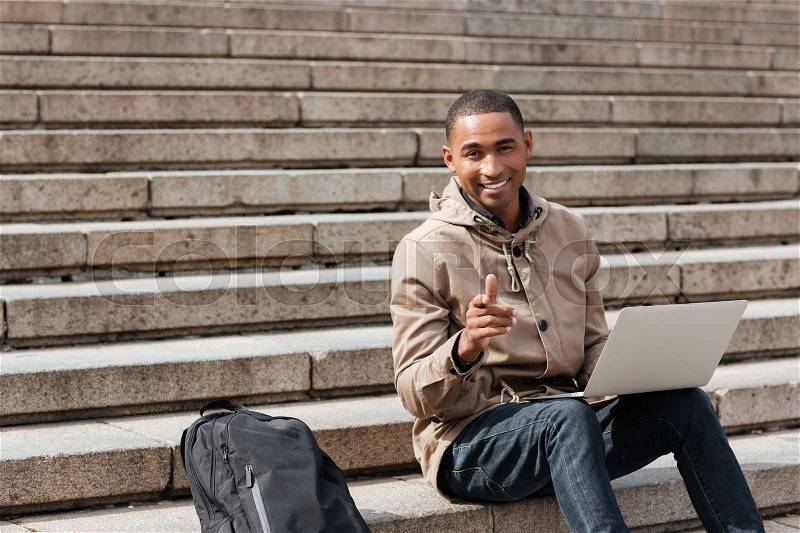Picture of young african happy man sitting on staircase and using laptop while pointing at camera. Looking at camera, stock photo