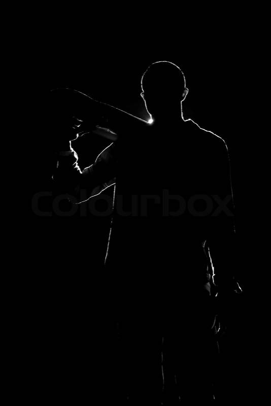 A backlit skateboarder guy posing under dramatic rear rim lighting with his skateboard in black and white, stock photo