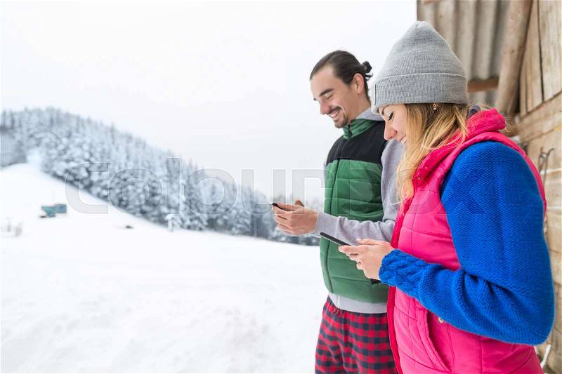 Young Couple Using Smart Phone Snowy Village Wooden Country House Man And Woman Online Messaging Winter Snow Resort Cottage Holiday Vacation, stock photo