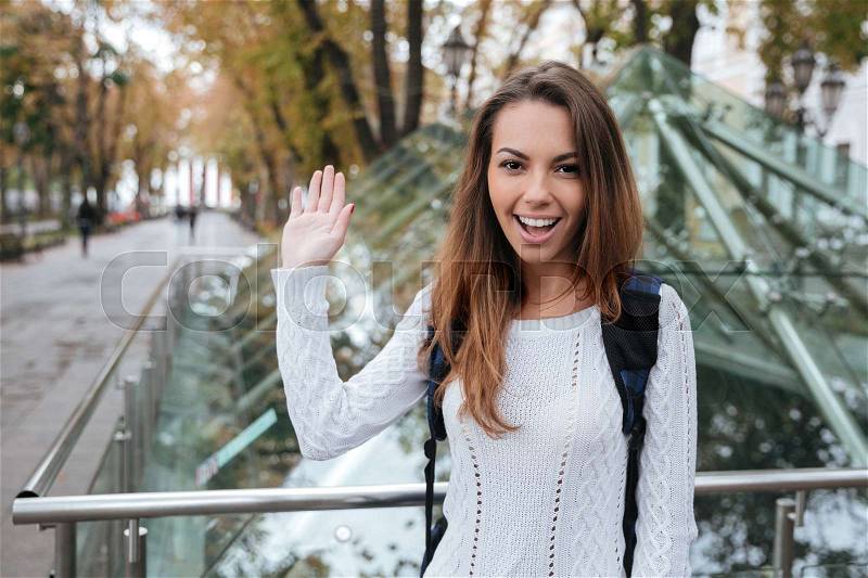 Smiling beautiful young woman waving and saying hello to you in park, stock photo