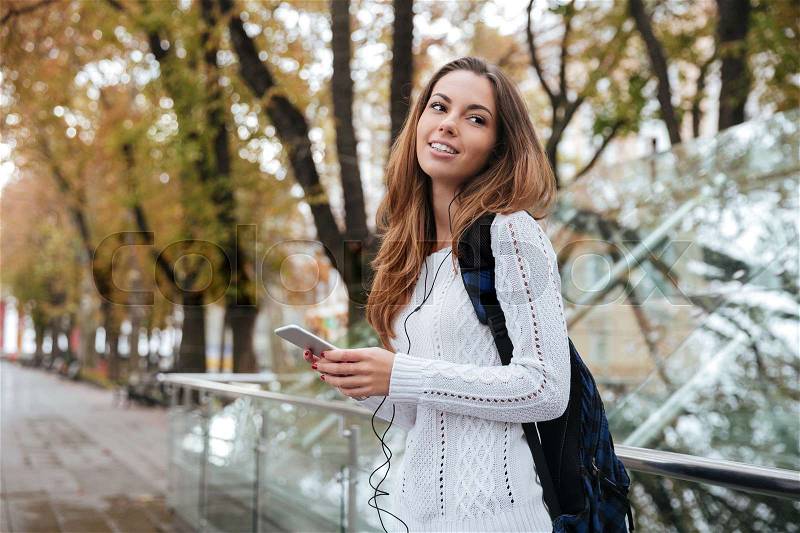 Smiling attractive young woman with backpack listening to music from cell phone in park, stock photo
