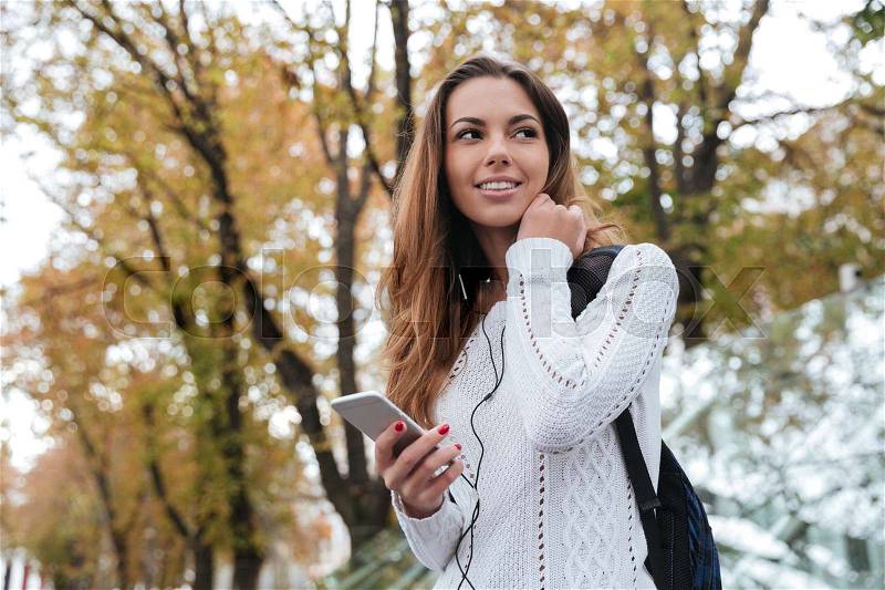 Happy charming young woman with earphones listening to music from cell phone outdoors, stock photo
