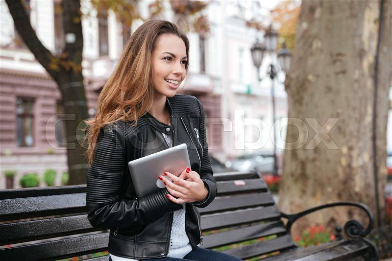 Smiling pretty young woman with tablet sitting on the bench in park, stock photo