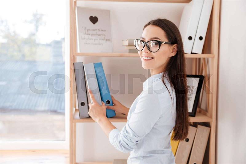 Cheerful young businesswoman taking folder from a book shelf while standing in office, stock photo