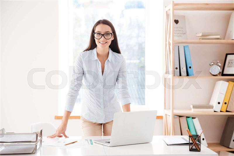 Young cheerful businesswoman standing at her office desk and looking at the camera, stock photo