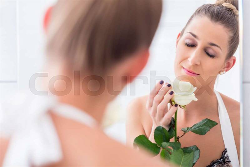 Happily smiling woman holding a white rose and regarding herself in the bathroom mirror, stock photo