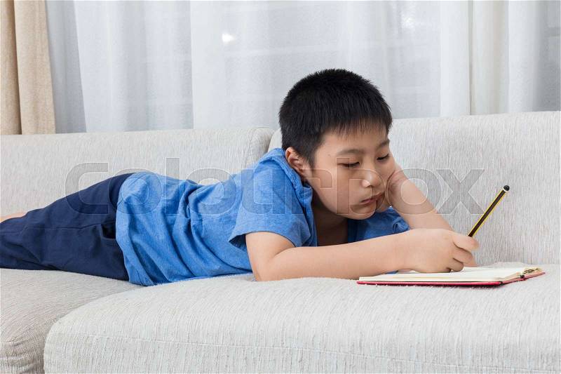 Bored Asian Chinese little boy writing book on the sofa in the living room at home, stock photo