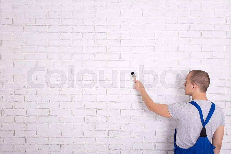 Back view of man painter in workwear painting white brick wall with brush and copy space, stock photo