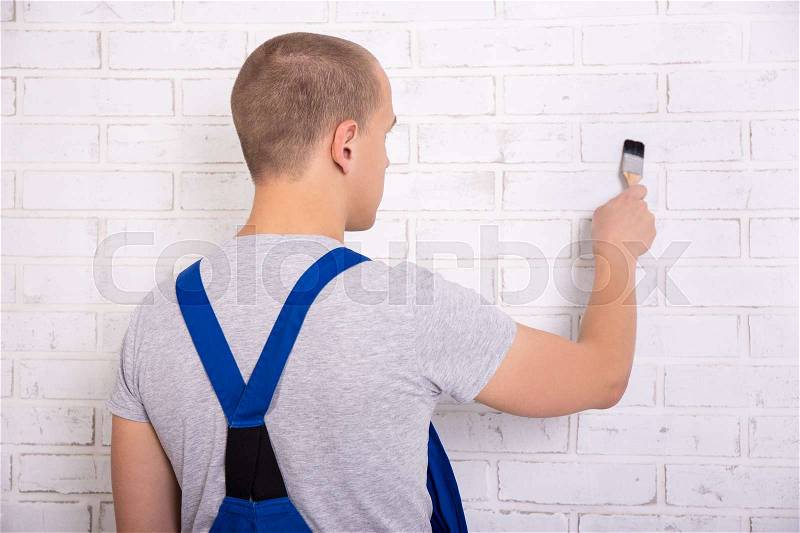 Back view of man painter in workwear painting white brick wall with brush, stock photo