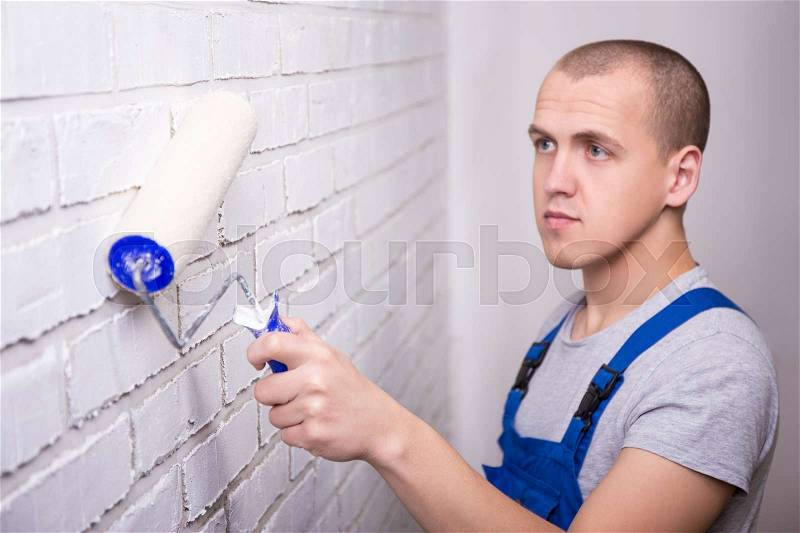 Handsome man painter in workwear painting white brick wall with paint roller, stock photo