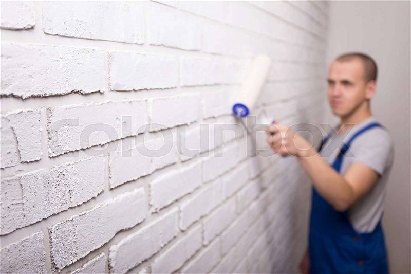 Young handsome man painter in uniform painting white brick wall with paint roller, stock photo