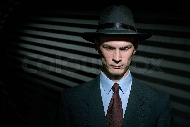 Fashionable young gentleman detective in suit and trilby hat with blinds in background and copy space, stock photo