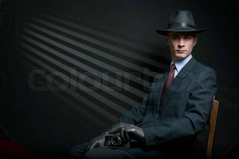 Stylish vintage detective posing on a chair in a fashionable hat , suit and gloves with his camera on his knee against a background of dark oblique parallel lines with copy space, stock photo