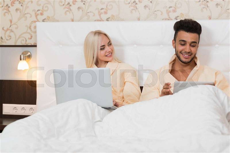Young Couple Lying In Hotel Bed, Happy Smile Hispanic Man And Woman Using Laptop And Tablet Computer, Lovers Bedroom, stock photo