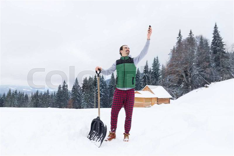 Young Man With Snow Shovel Using Smart Phone Chatting Online Village Wooden House Winter Resort Cottage Holiday Vacation, stock photo
