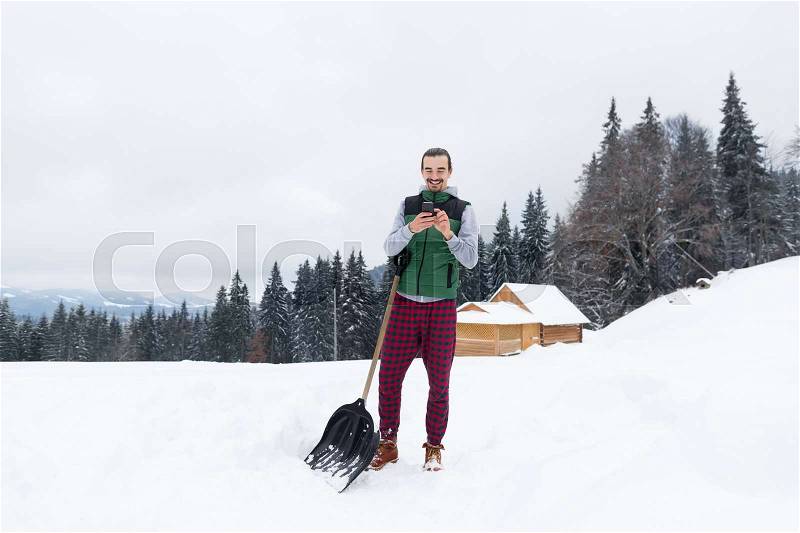 Young Man With Snow Shovel Using Smart Phone Chatting Online Village Wooden House Winter Resort Cottage Holiday Vacation, stock photo