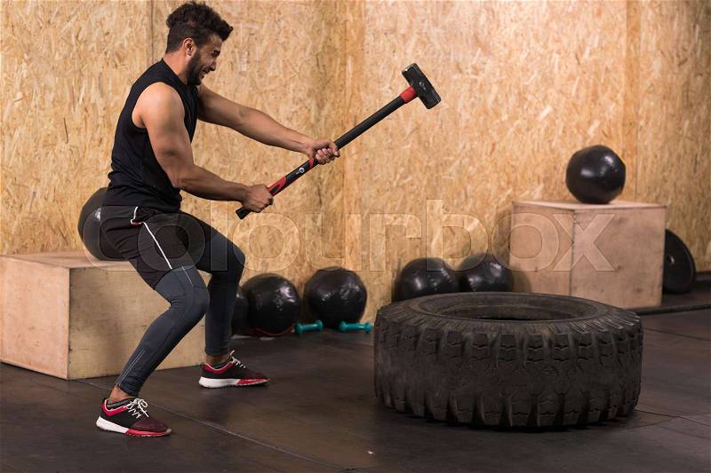 Sport Fitness Man Hitting Wheel Tire With Hammer Sledge Crossfit Training, Young Healthy Guy Gym Interior, stock photo
