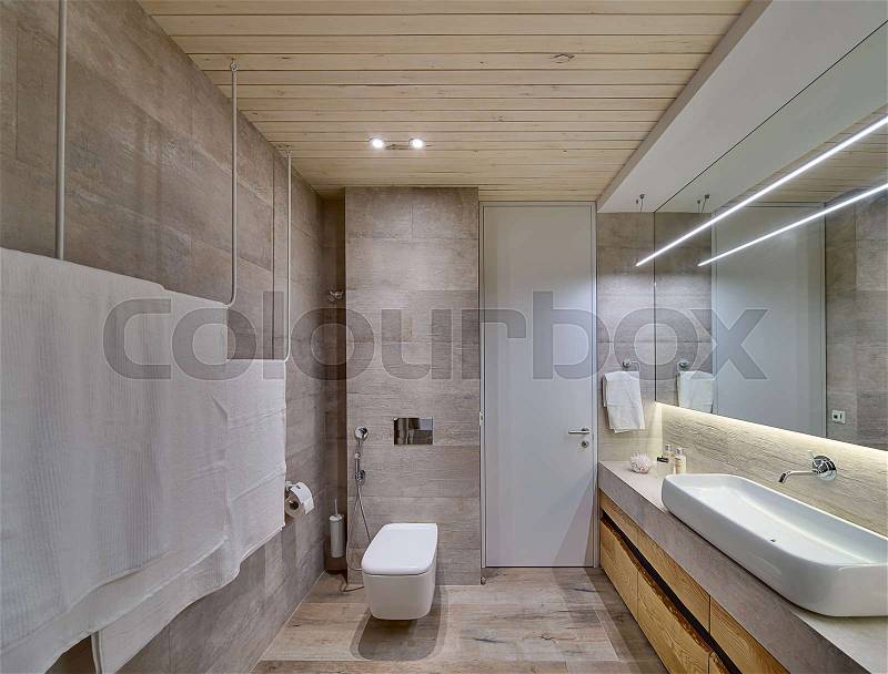 Bathroom in a modern style with textured tiles and a wooden ceiling. There is a white sink on the rack, wooden lockers, mirror, towel holders, toilet, shower, door, wooden panel above the sink, lamps, stock photo