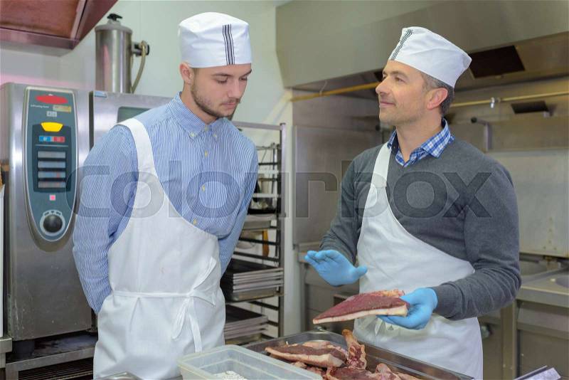 Apprentice and chief preparing meat in restaurant kitchen, stock photo