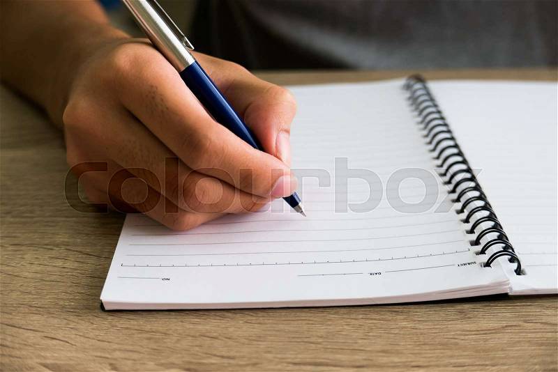 Closeup of male right hand writing text on blank notebook on wooden desk in office, stock photo