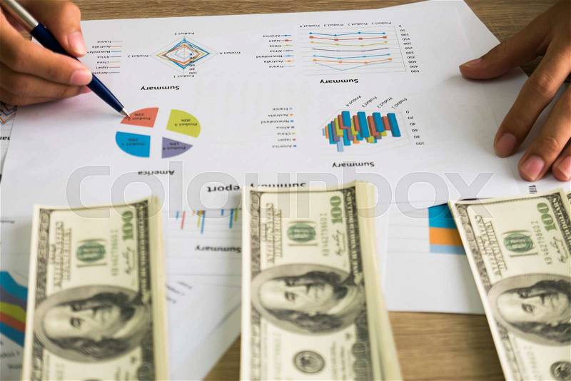 Closeup of Asian male right hand hold ball pen points on business charts under bunch of dollars banknotes for business, finance, tax and people concept, stock photo