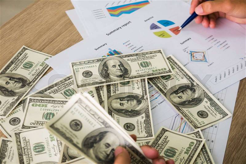 Closeup of Asian male left hand counting bunch of dollars banknotes and his right hand hold ball pen points on business charts background for business, finance, tax and people concept, stock photo
