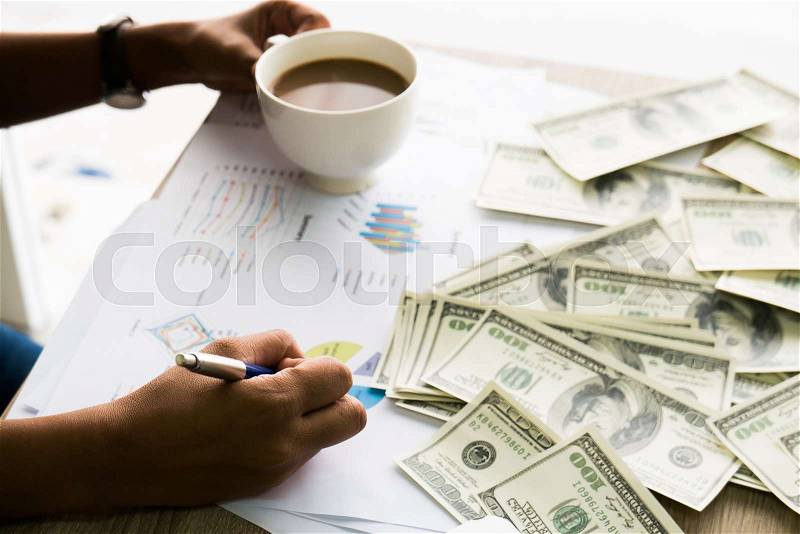 Closeup of Asian female writes something on business charts under bunch of dollars banknotes nearby her labtop and a cop of coffee for business, finance, tax and people concept, stock photo