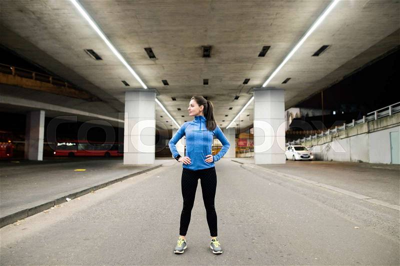 Beautiful young runner under the bridge in the town in the evening resting, arms on hips, stock photo