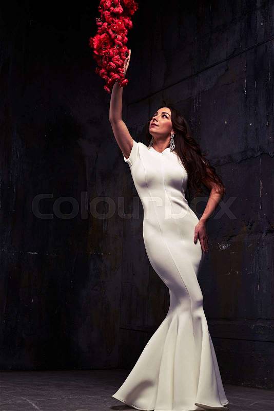 Full-length portrait of stunning woman in white evening dress. Touching a swag of red roses. Isolated in studio. Looking upwards, stock photo